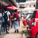People walking along a busy sidewalk in central Paramaribo, Suriname, with parked cars and storefronts.