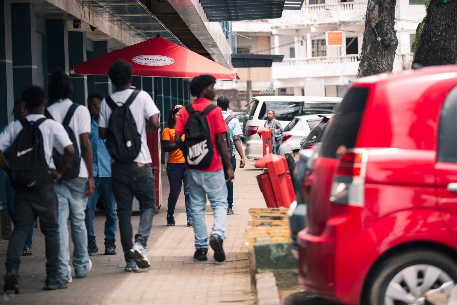 People walking along a busy sidewalk in central Paramaribo, Suriname, with parked cars and storefronts.