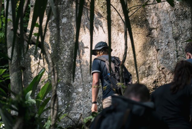 Hiker walking along a narrow rainforest trail beside a massive granite rock formation during the Voltzberg expedition in Suriname.