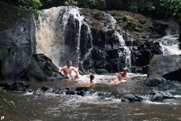 Travellers swimming in a natural pool beneath a cascading waterfall surrounded by dark granite rocks and tropical rainforest in Suriname.