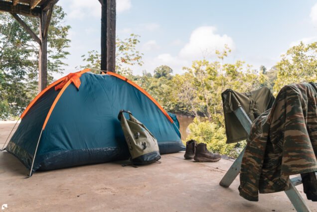 A blue and orange camping tent beneath a wooden shelter beside the rainforest and river at Raleigh Falls, Suriname.